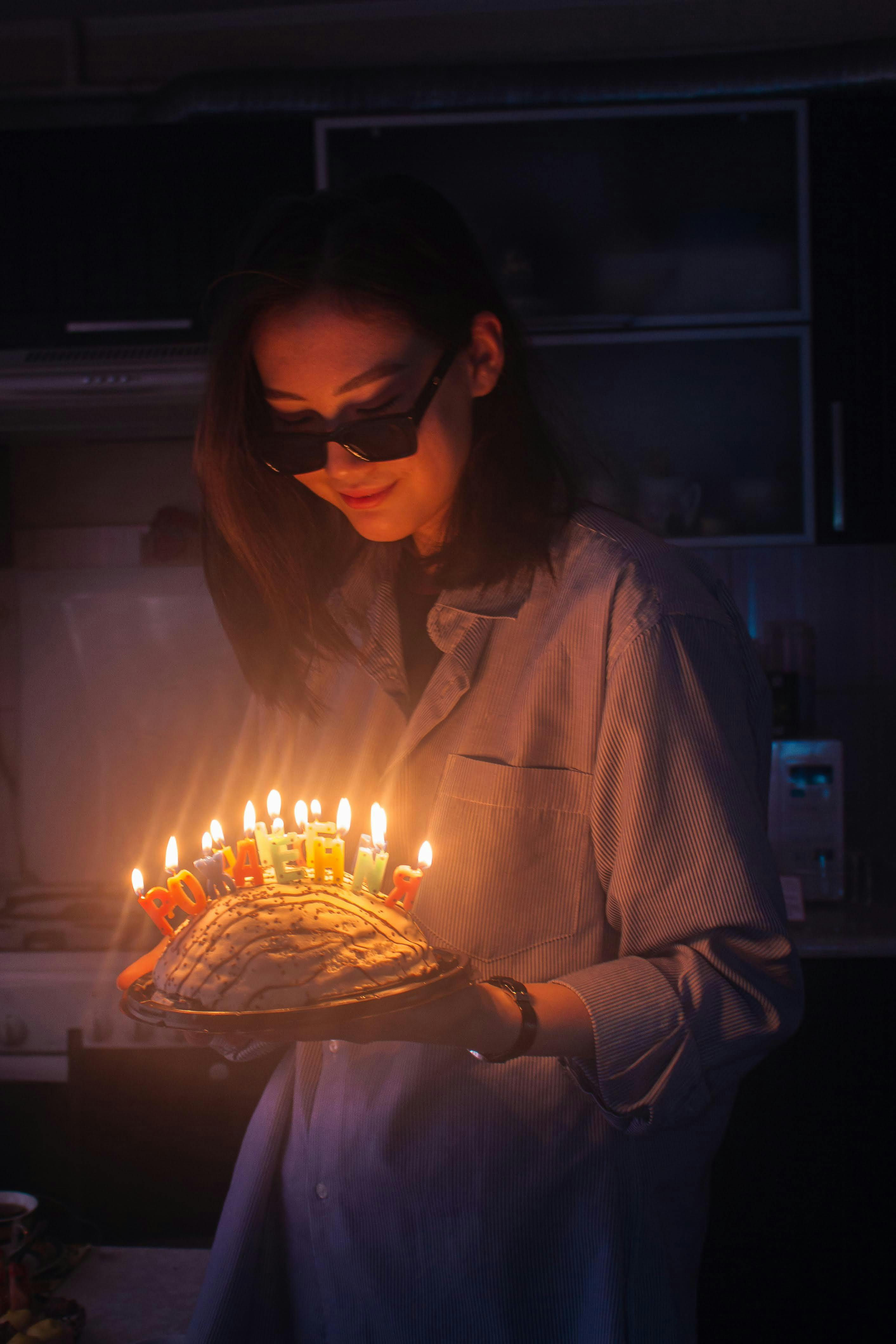 A young woman wearing sunglasses holds a cake with lit candles in a dark room, capturing a birthday celebration.