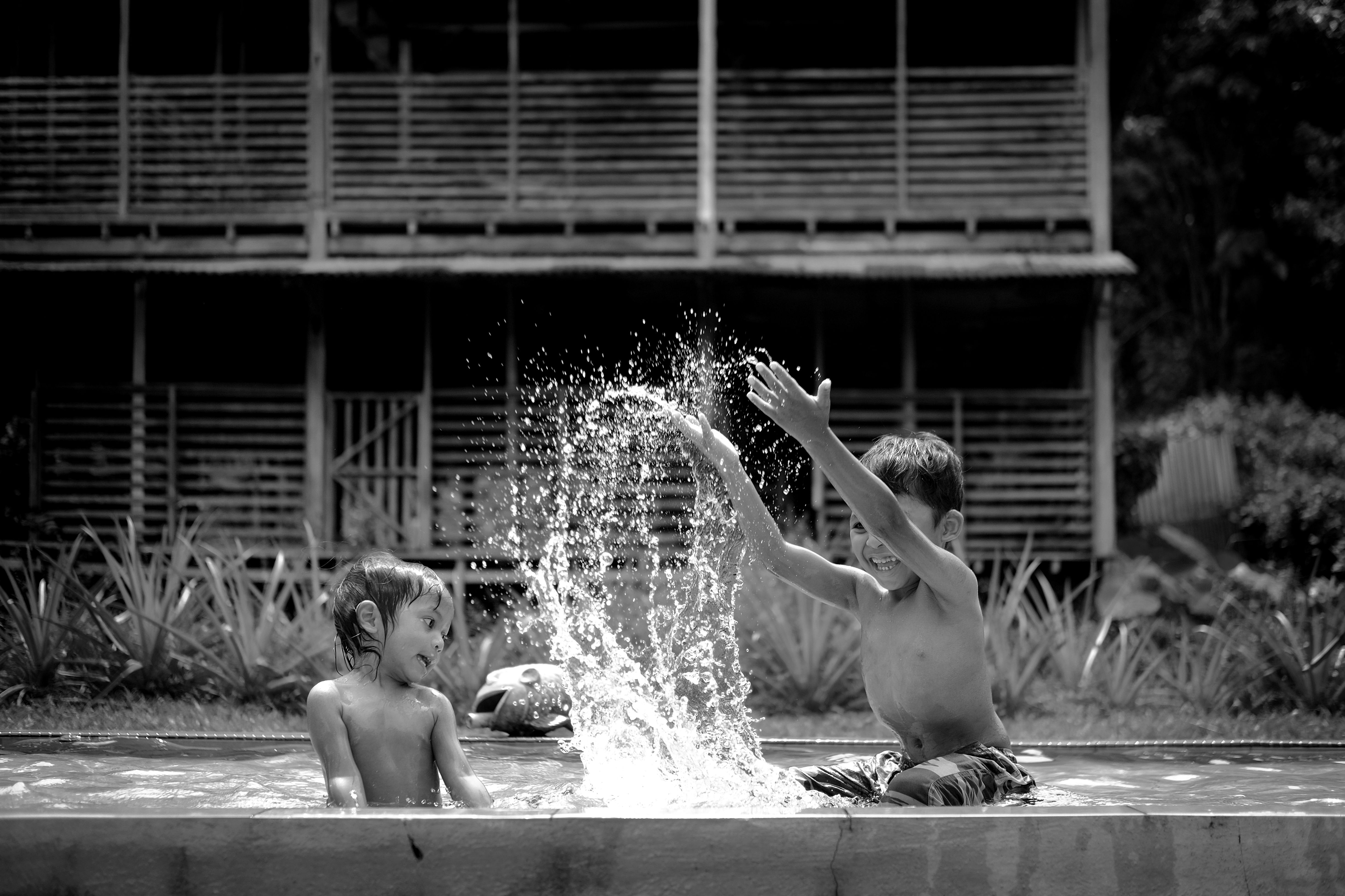 Two children enjoying a fun moment splashing water in an outdoor pool. Black and white photography.