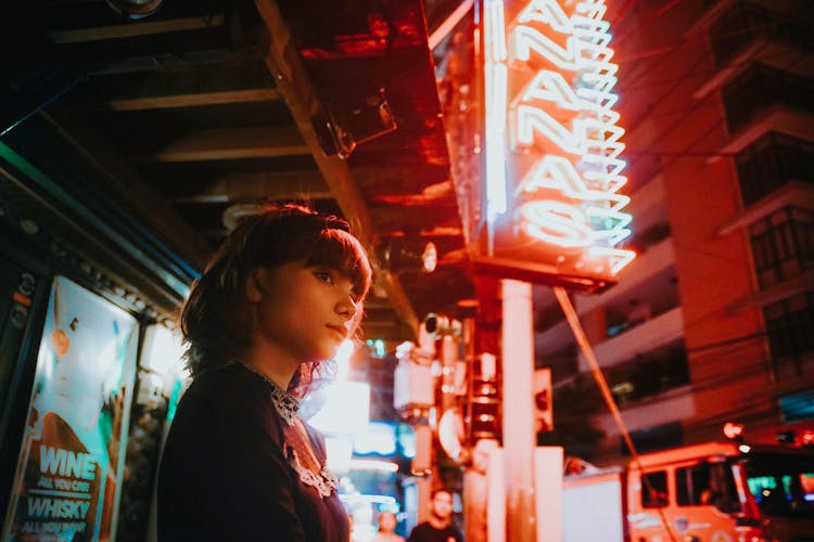 Young Woman Standing On The Sidewalk Under A Neon Sign At Night