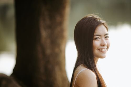 Free A young Asian woman with long hair smiling beside a tree in daylight. Stock Photo