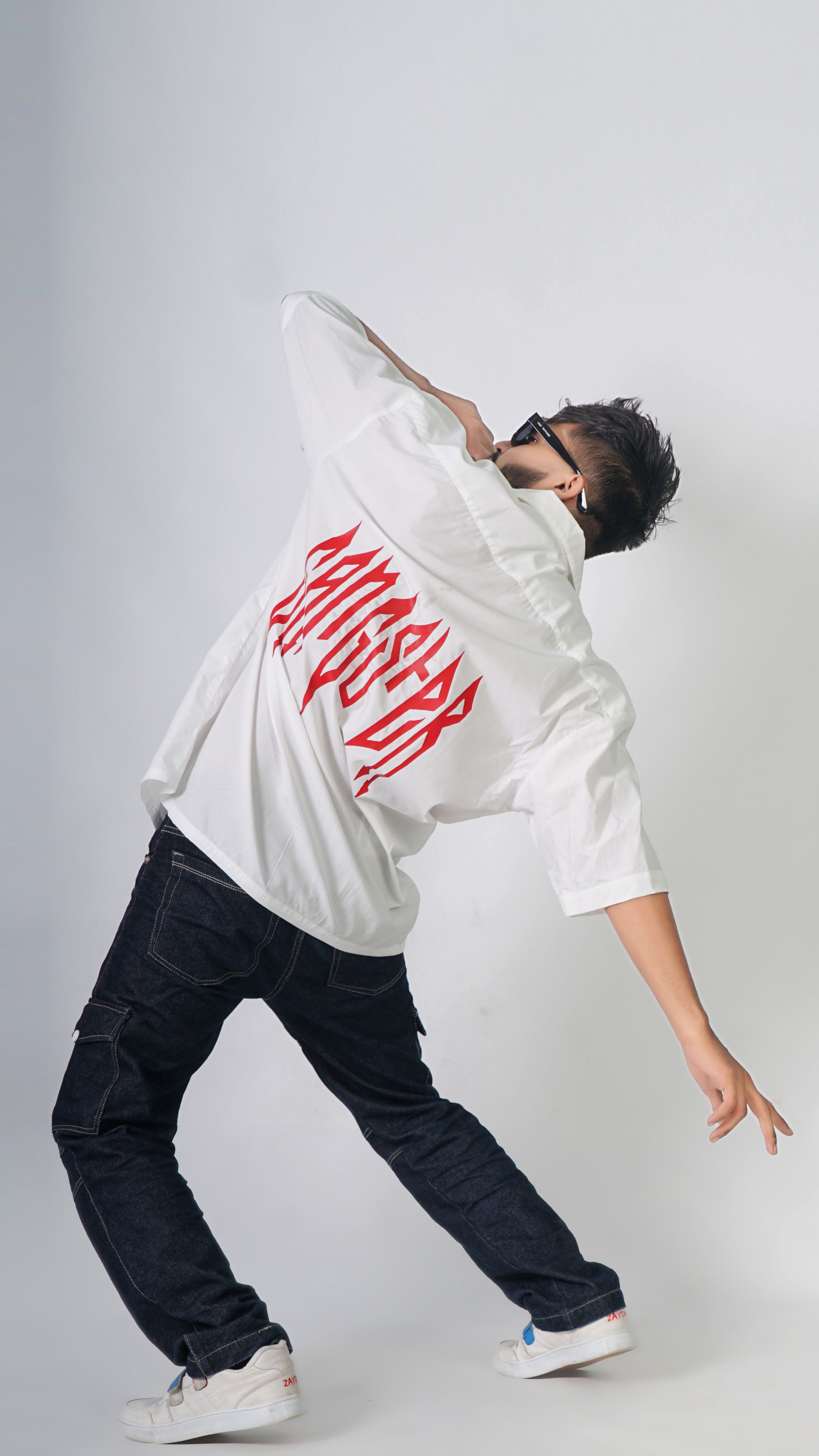 Model in sunglasses poses dynamically with a white printed shirt in a studio fashion shoot.