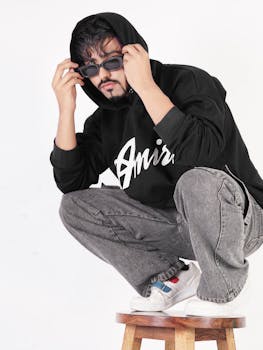 Stylish man in a black hoodie and sunglasses posing on a wooden stool in studio.