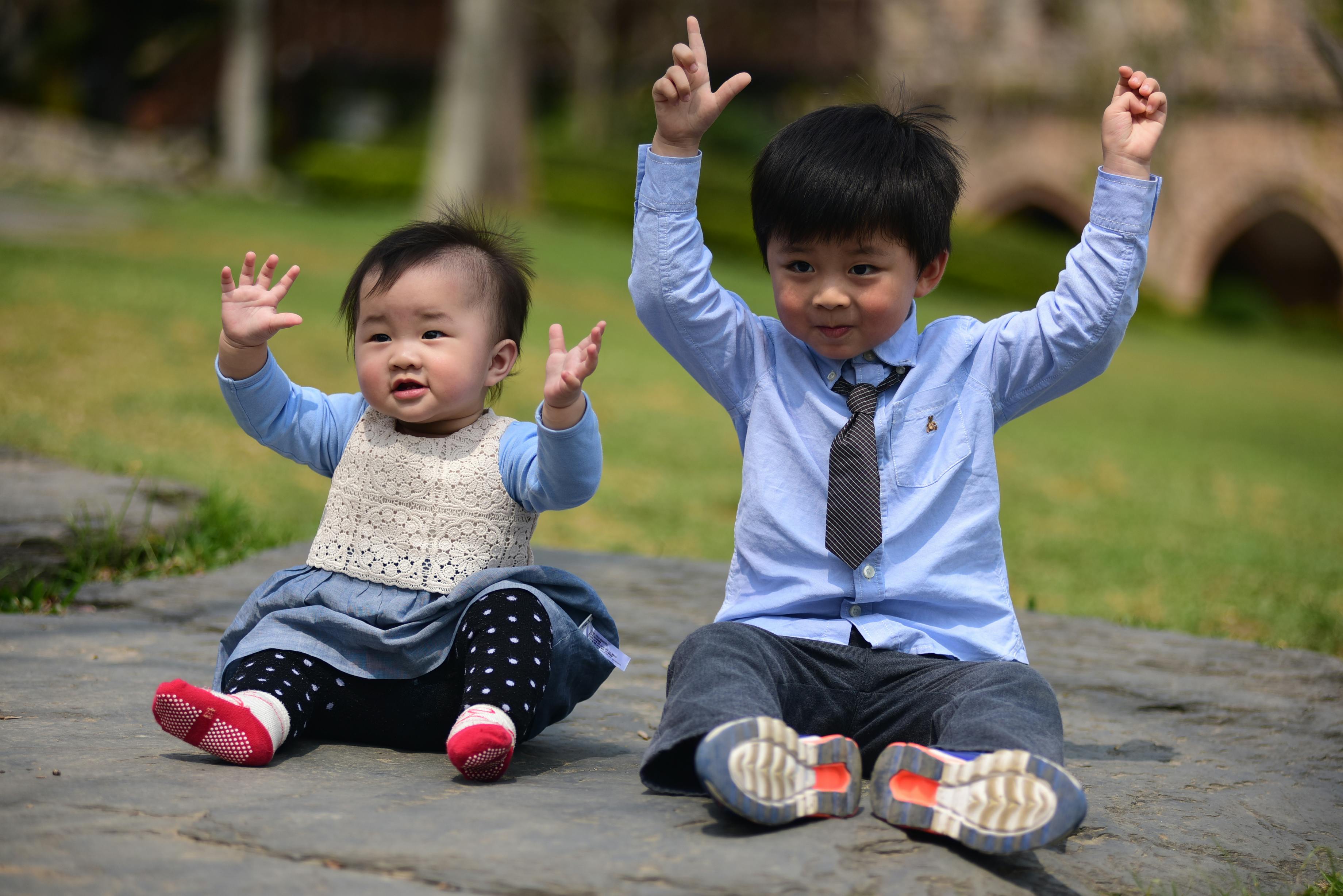 Two happy young children sitting on the ground with arms up, enjoying a sunny day outdoors.