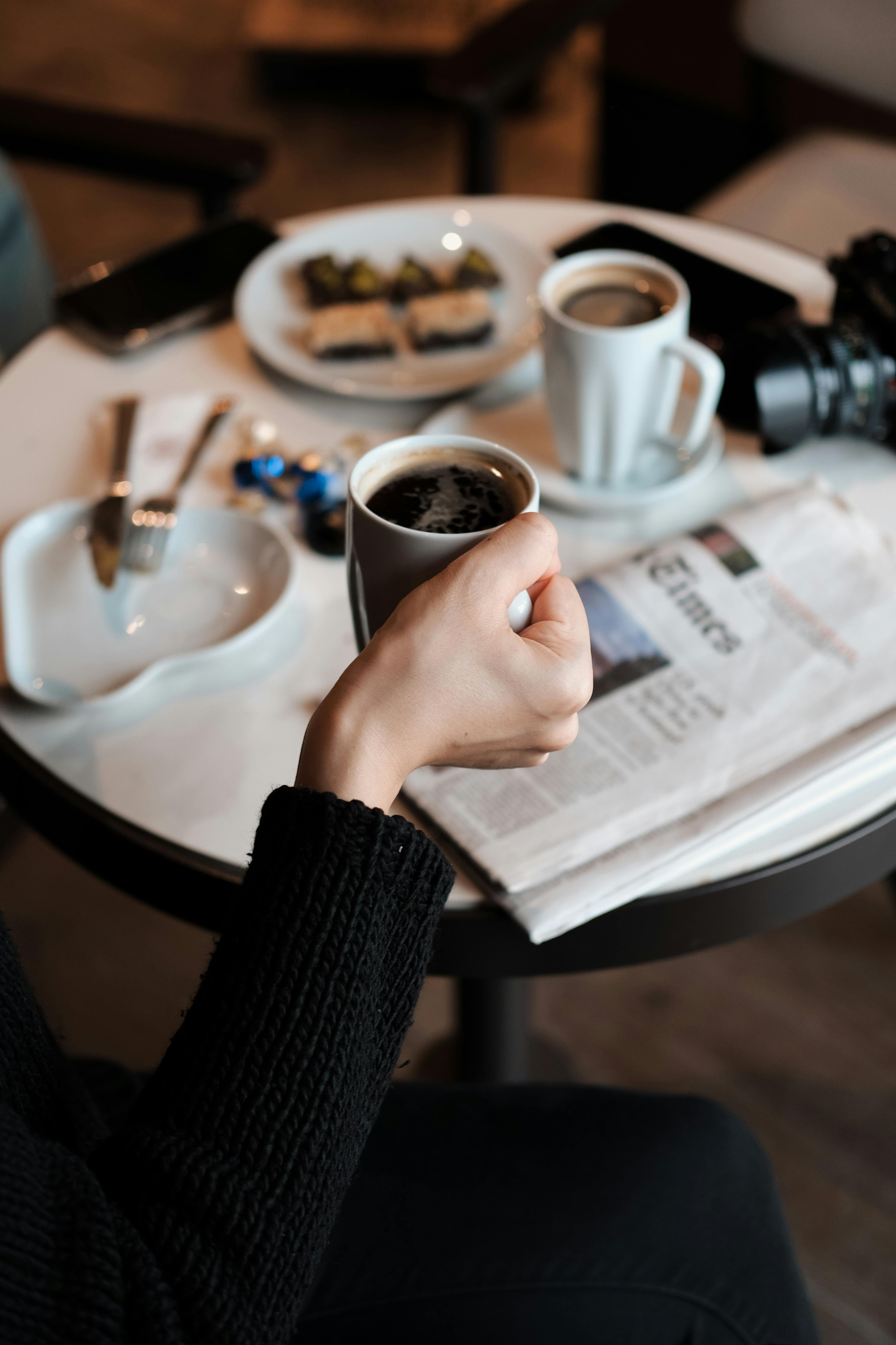 A cozy scene with a hand holding a coffee cup on a table with snacks and a newspaper.