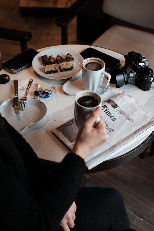 Free Person holding coffee cup at cafe table with newspaper, camera, and pastries. Stock Photo