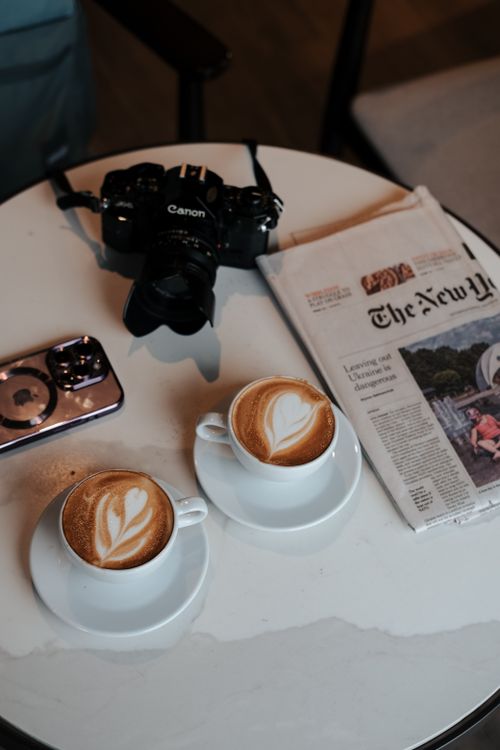 Free An inviting scene with cappuccino, camera, and newspaper on a table. Stock Photo