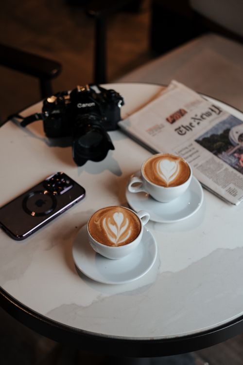 Free Two cappuccinos with latte art on a table with a camera and newspaper, ideal for lifestyle themes. Stock Photo