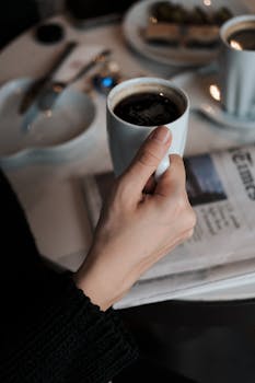 A hand holding a coffee cup over a newspaper in a cozy café setting. Calm and inviting.