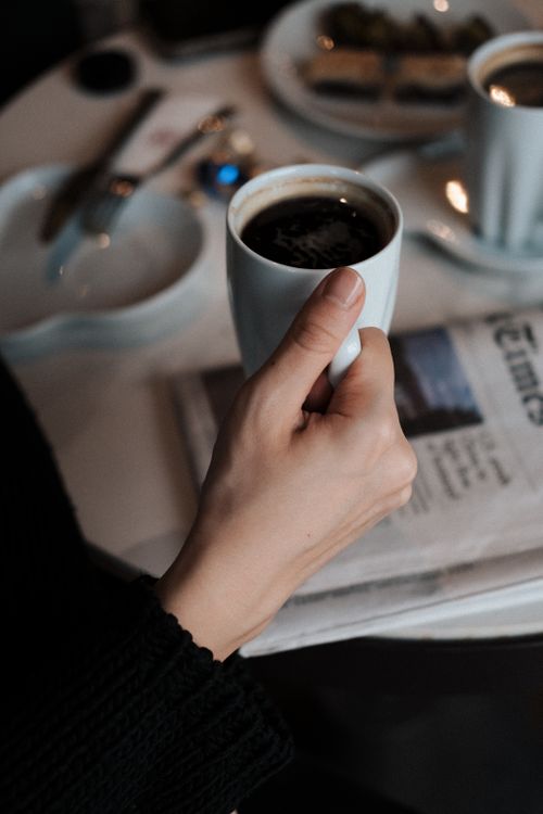 Free A hand holding a coffee cup over a newspaper in a cozy café setting. Calm and inviting. Stock Photo