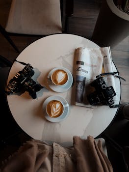 Overhead view of cappuccinos and cameras on a cafe table with newspaper.