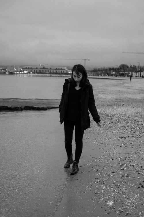 Free Black and white photo of a woman walking along the beach at twilight with city lights in the background. Stock Photo