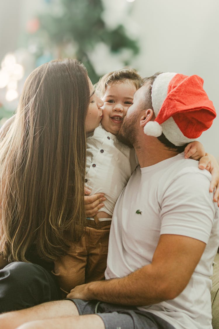 Mother And Father Kissing Their Baby On The Cheeks In Front Of A Christmas Tree