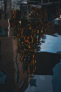 A blurred reflection of a decorated Christmas tree with lights on an urban water surface.