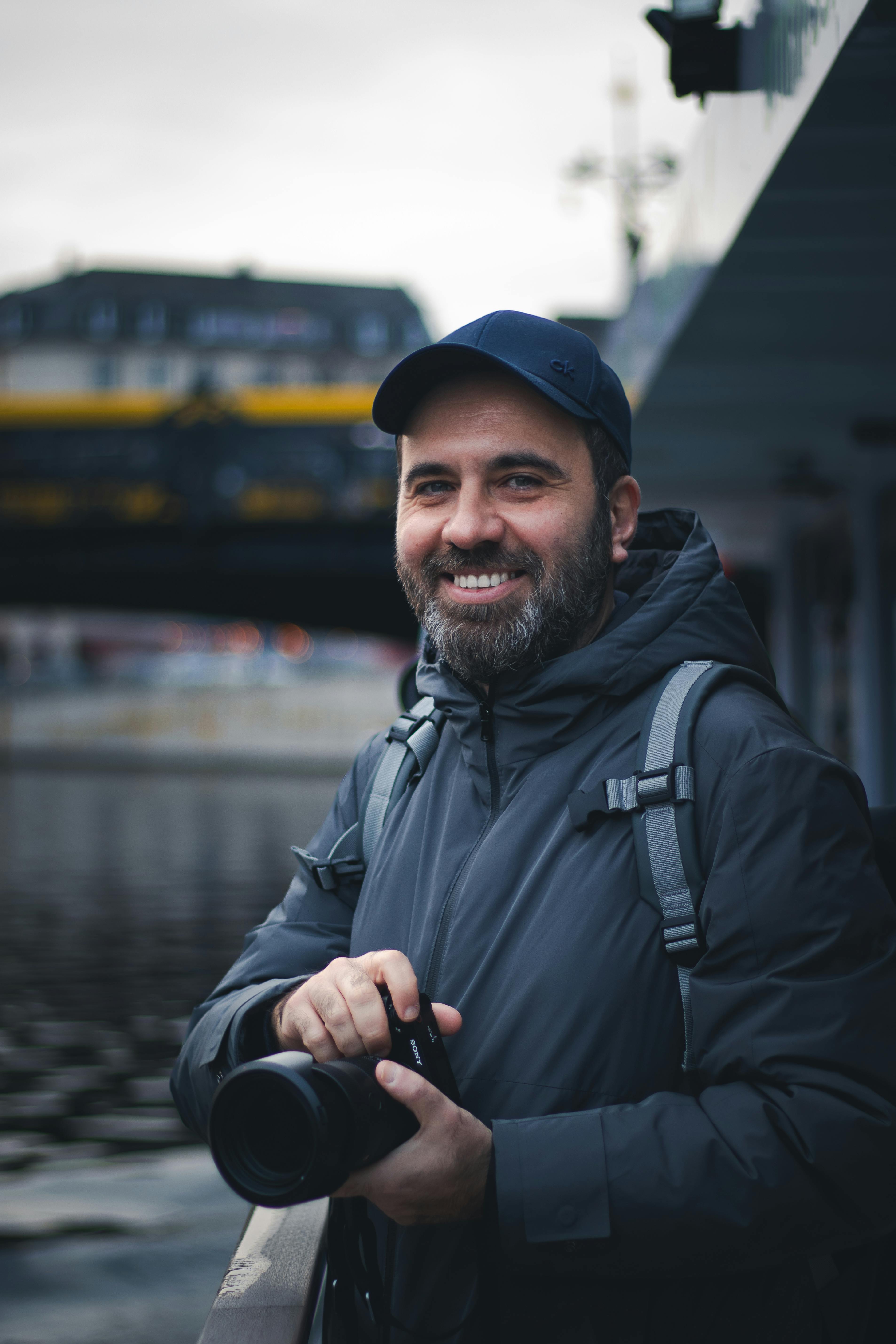 Man with a camera smiling by a river bridge in fall setting. Perfect for lifestyle photography themes.
