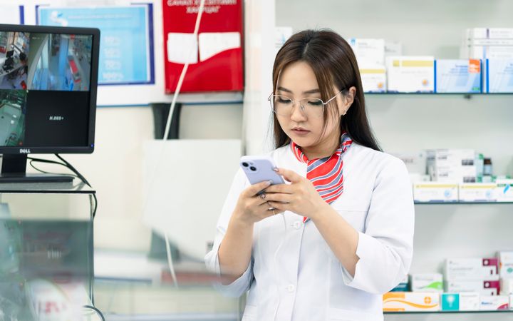 A female pharmacist in a lab coat using a smartphone in a pharmacy, surrounded by medicine shelves.
