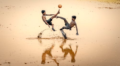 Boys Jump Kicking a Ball Over a Muddy Water