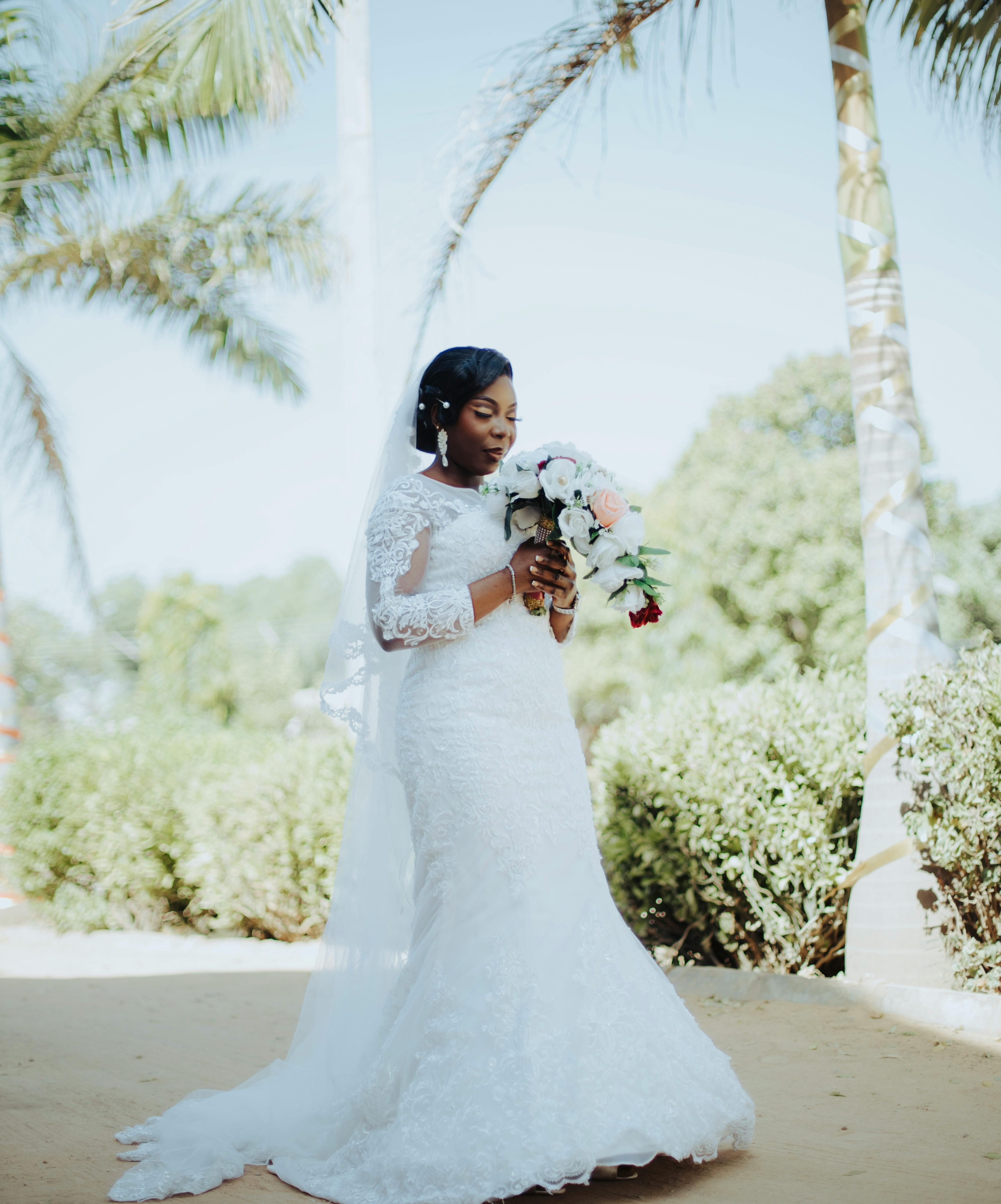 Bride in lace wedding dress holding bouquet outdoors under palm trees.
