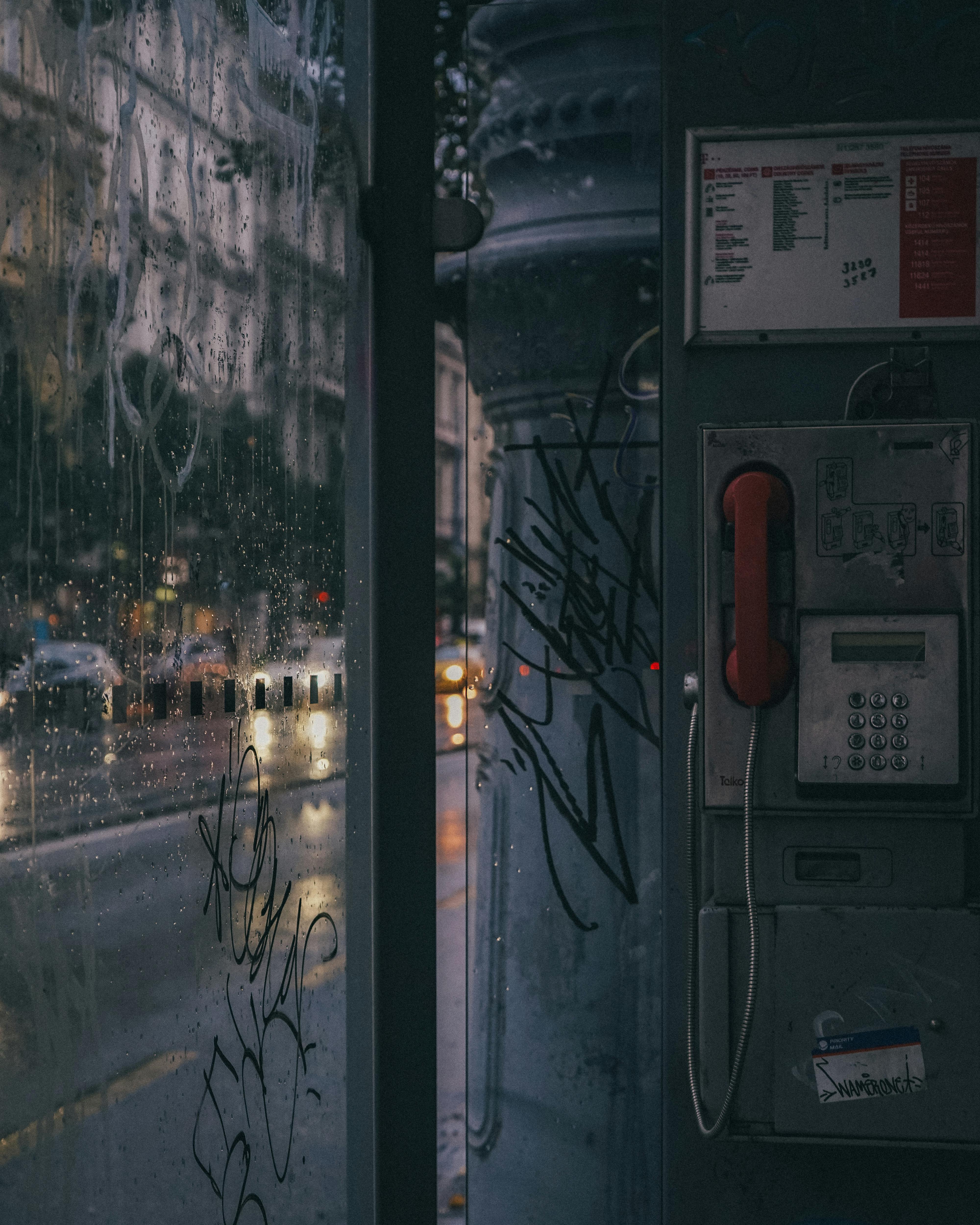 A vintage phone booth with graffiti in a rainy urban Budapest street.