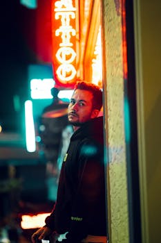 A man in a black hoodie stands against an urban wall under vibrant neon lights at night.