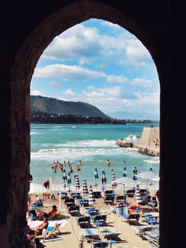 Picturesque view of Cefalù beach through an arch, capturing vacationers and the serene sea.