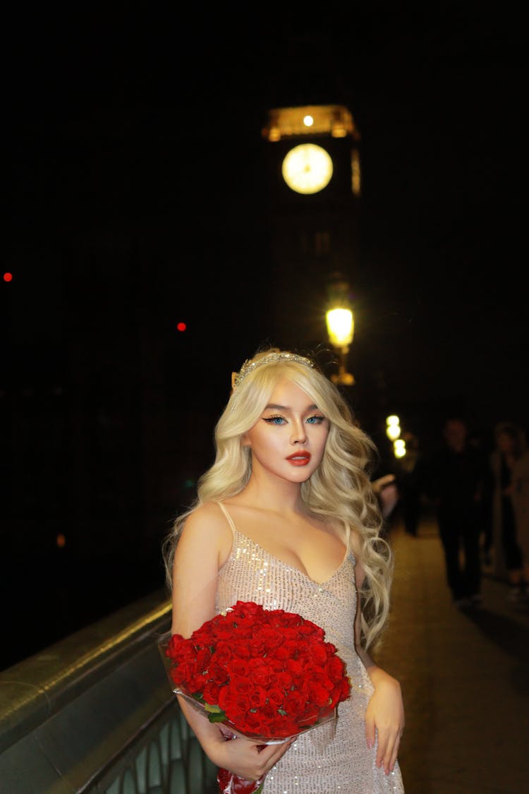 Young Woman In A Dress Standing Outside At Night And Holding A Bouquet Of Red Roses 