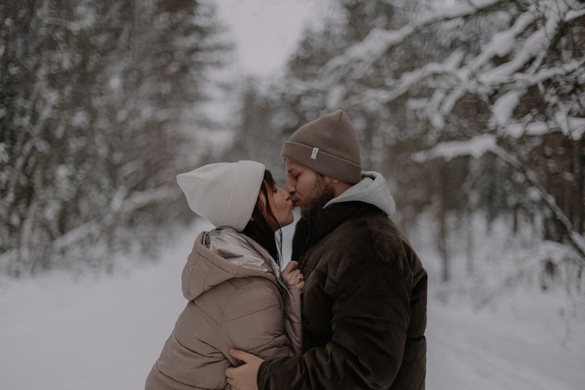 A couple shares a romantic kiss on a snowy forest path during winter, surrounded by conifer trees.