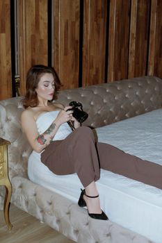 A fashionable woman with tattoos relaxes on a plush bed, examining her camera.