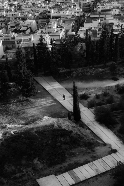 Black and white aerial view of a park pathway cutting through an urban cityscape, featuring trees and a solitary person.