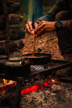 Person cooking with a pan and kettles on an open fire in the forest, focusing on hands.