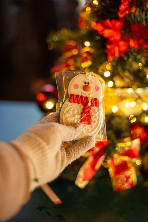 Free Gloved hand holding a wrapped snowman cookie in front of a decorated Christmas tree. Stock Photo