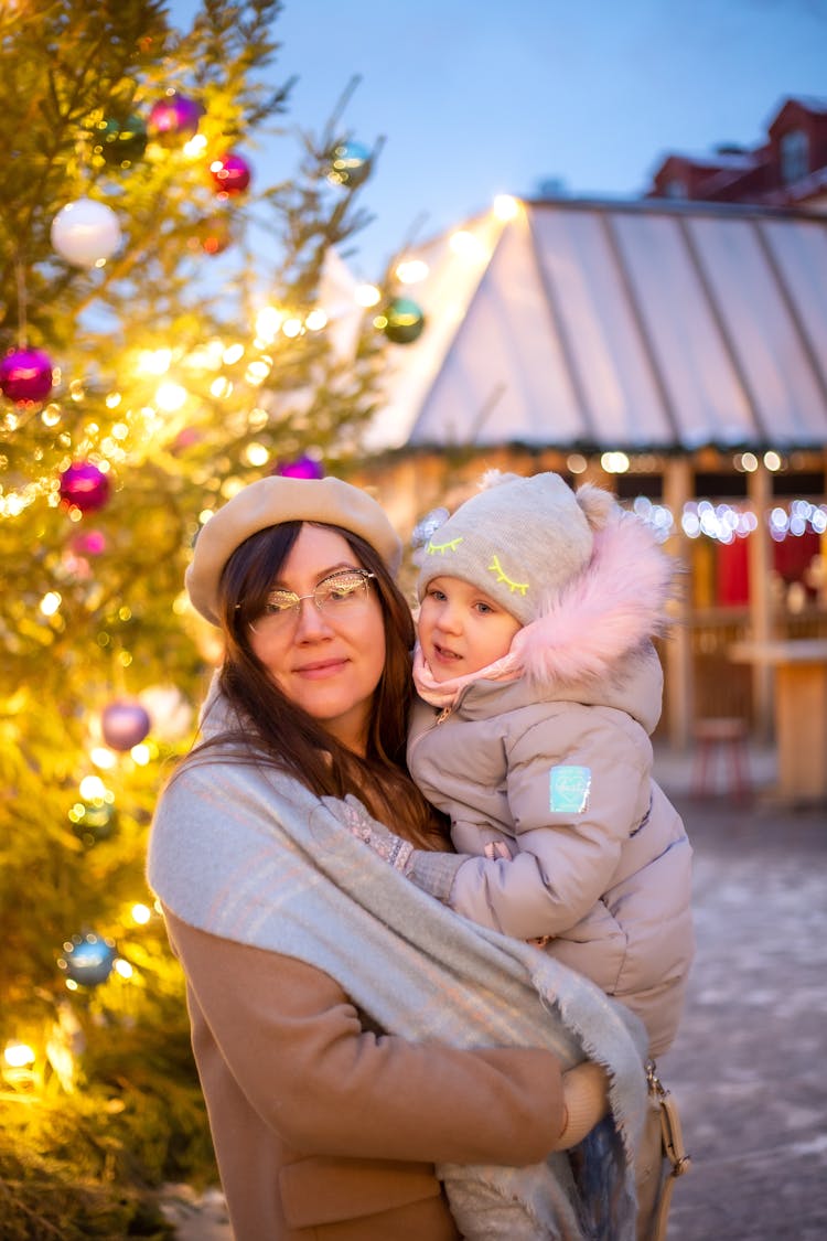 Mother Holding A Child And Standing Next To A Christmas Tree Outside 