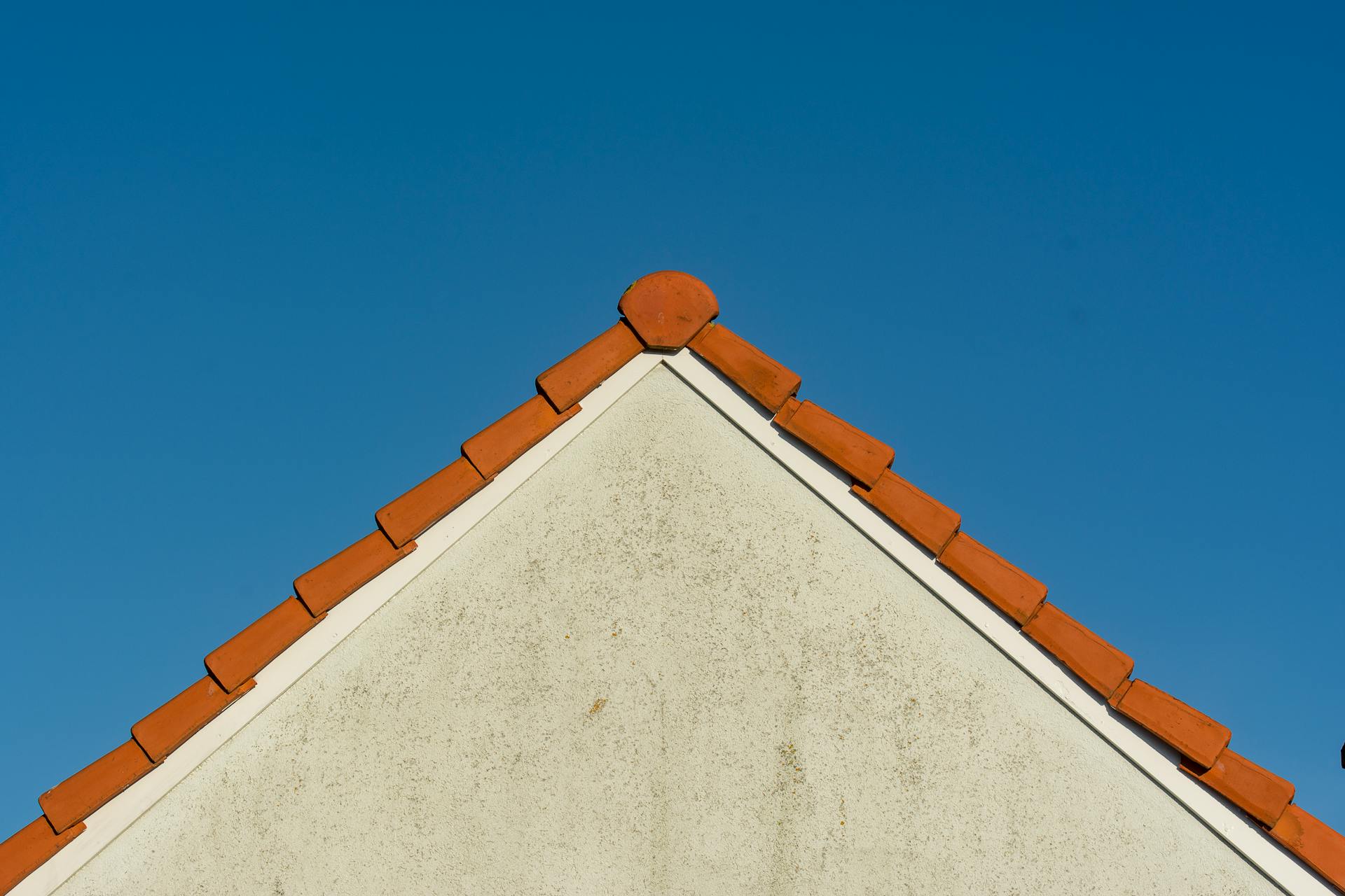 Minimalist image of a gable roof with red tiles against a clear blue sky.