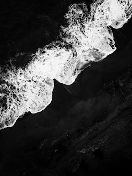 A dramatic black and white aerial shot of waves crashing on a dark shore.