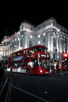 Brightly lit London street at night featuring a classic red double-decker bus and historic architecture.