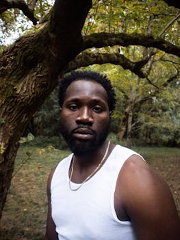 A thoughtful portrait of a man standing in a forest, wearing a white tank top.
