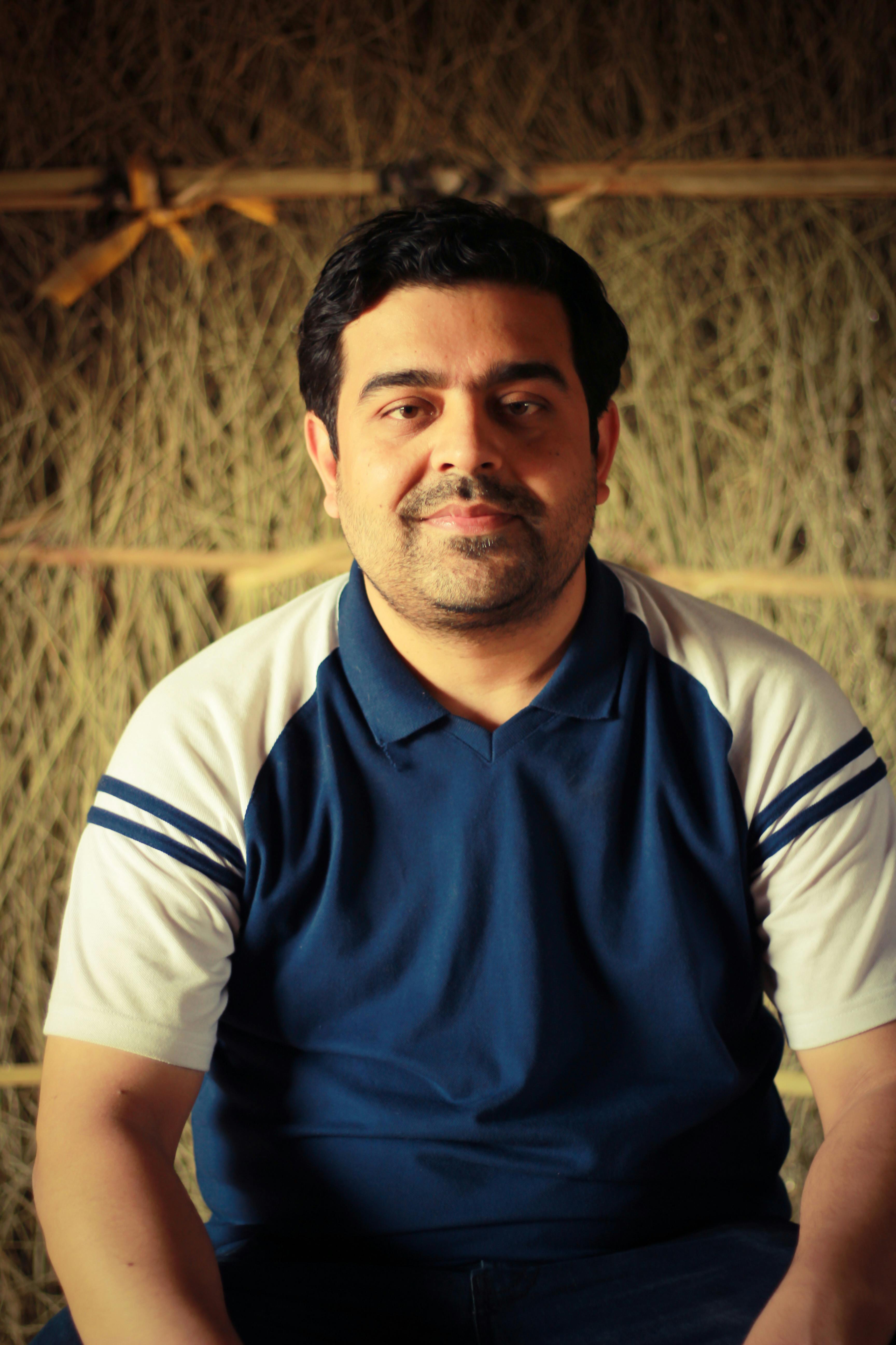 Man posing in front of hay bales, wearing a blue and white T-shirt with a thoughtful expression.