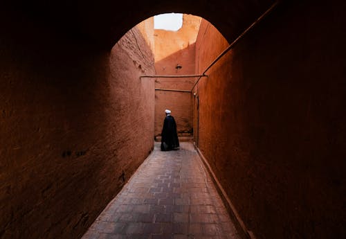 Person in a Black Gown Walking in a Narrow Alley between Buildings 
