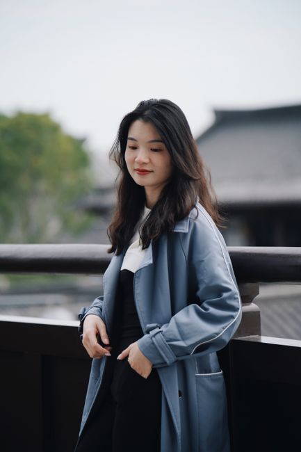 Stylish young woman in a blue coat posing on a terrace with a serene expression, embracing modern fashion.