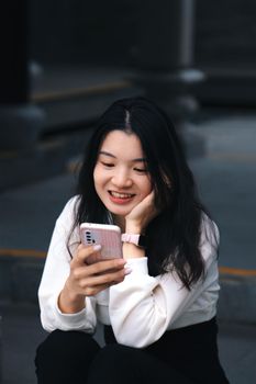 A young woman sitting outside, smiling while looking at her smartphone.