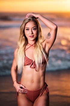 A young woman in a bikini poses on the beach at sunset, showcasing summer fashion.