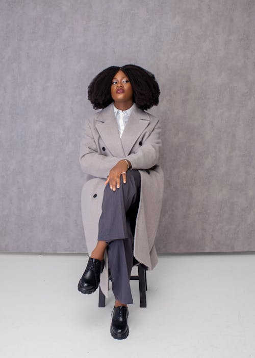Photo of a Woman with Afro Hairstyle, Wearing a Gray Coat, Posing against a Gray Wall