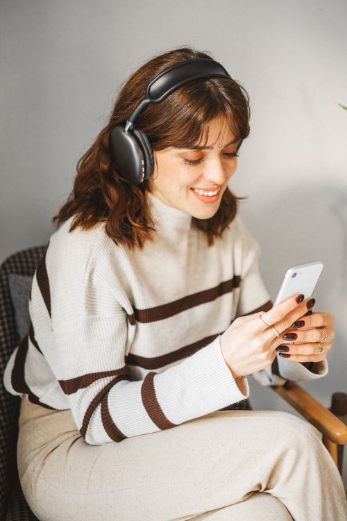 Free Woman wearing wireless headphones, seated indoors, browsing on smartphone. Stock Photo