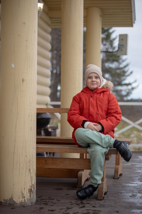 Free A child in a red jacket sits on a wooden bench in an outdoor terrace setting, surrounded by pillars. Stock Photo