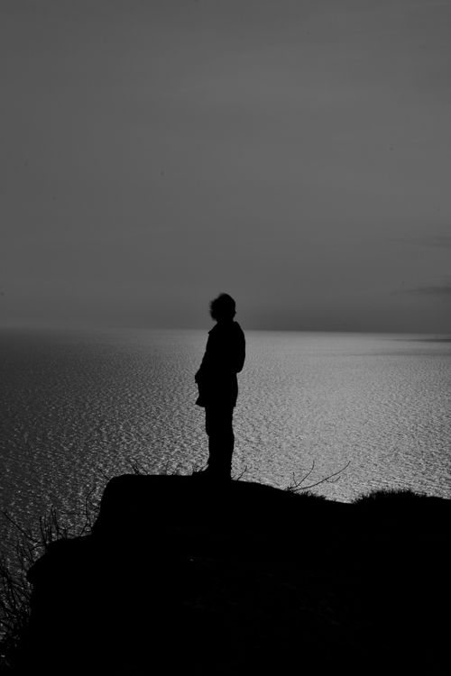 Free A person stands silhouetted on a cliff, gazing at the tranquil sea under a twilight sky. Stock Photo