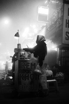 Black and white image capturing a street vendor at night with fog, creating a moody urban atmosphere.