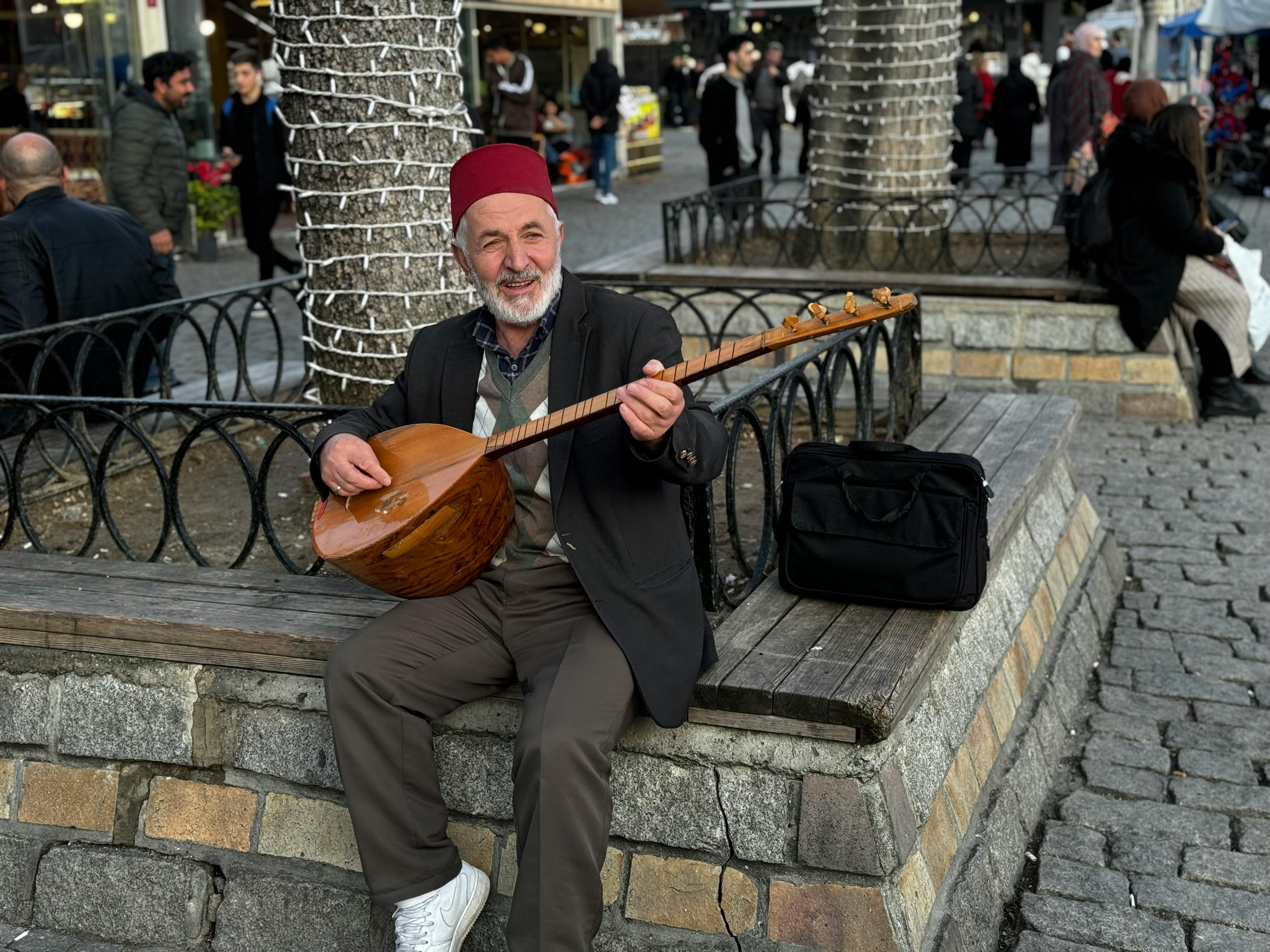Elderly Man in a Fez Hat Playing a Traditional Baglama Lute Sitting on the City Walkway