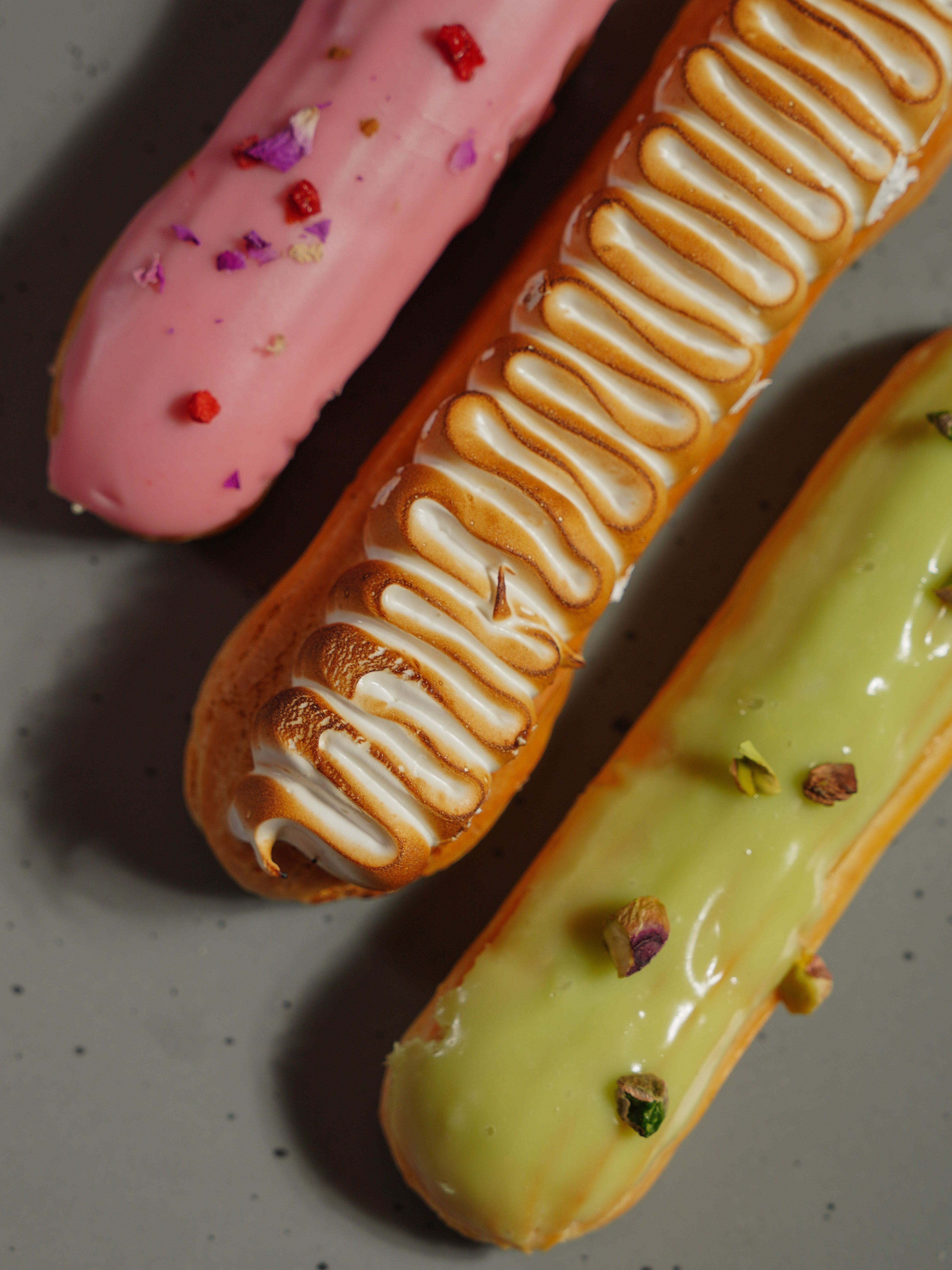Close-up of three colorful eclairs with unique toppings on a gray surface.