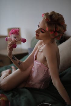 A stylish woman in a pink corset holds a toy pony indoors, relaxing with retro curls.