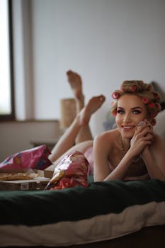 Smiling woman with hair curlers enjoying a relaxed day in a bedroom setting, surrounded by snacks.
