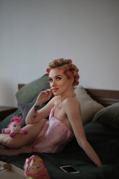 A stylish woman in pink rollers and lingerie relaxing with a toy unicorn on a bed.